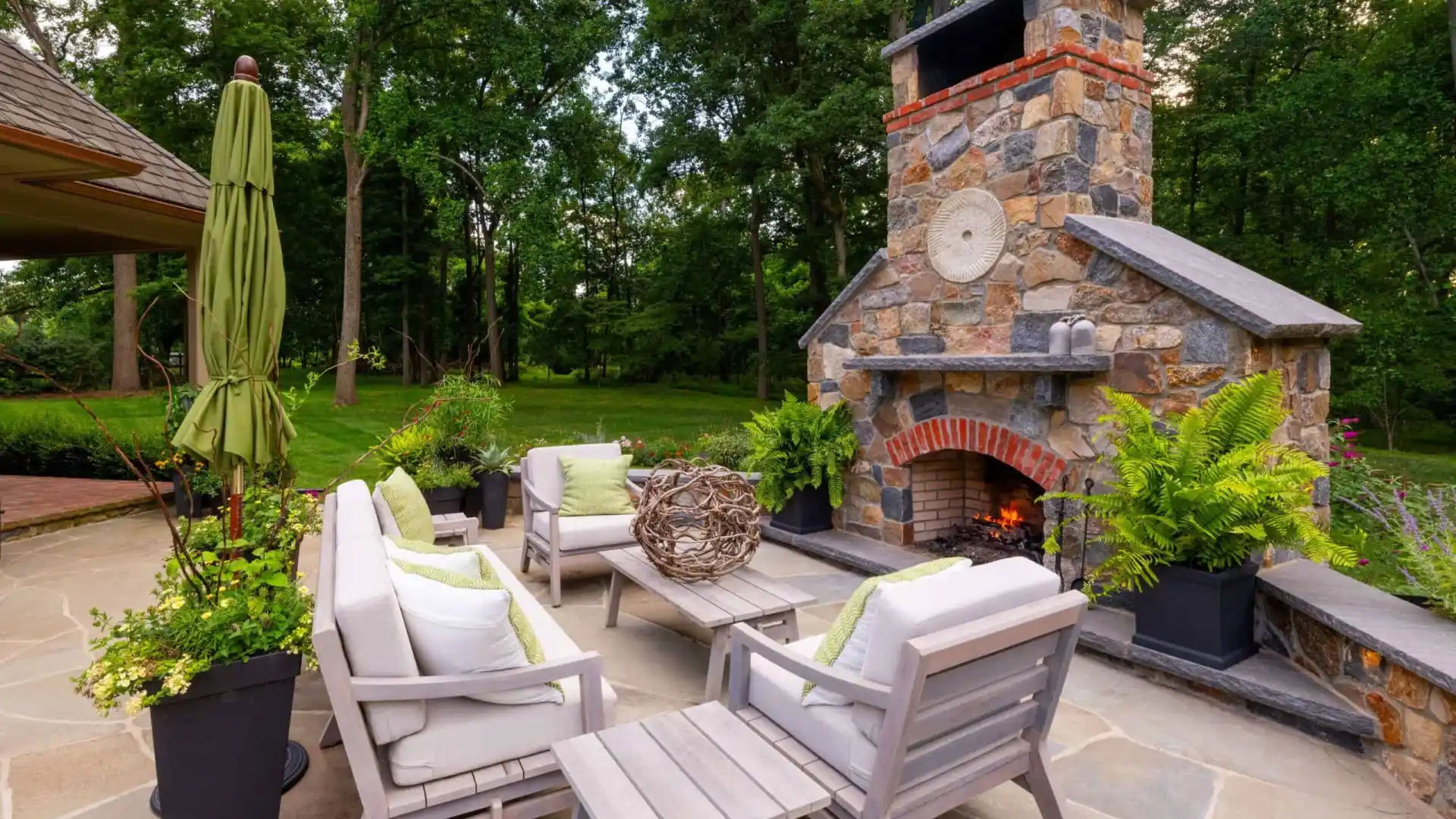Outdoor patio with stone fireplace, surrounded by wooden chairs and a coffee table adorned with decorative twigs. Ferns and potted plants accent the space, crafted by top landscaping services in Delaware County PA. A green umbrella shades to the left, framed by lush trees.