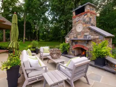 Outdoor patio with stone fireplace, surrounded by wooden chairs and a coffee table adorned with decorative twigs. Ferns and potted plants accent the space, crafted by top landscaping services in Delaware County PA. A green umbrella shades to the left, framed by lush trees.