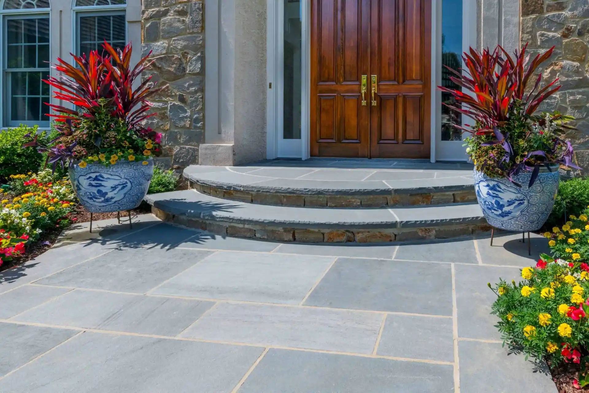 Elegant stone porch with curved steps leading to double wooden doors, flanked by large blue-and-white planters with vibrant red and green foliage. Colorful flowers border the slate-tiled entryway.