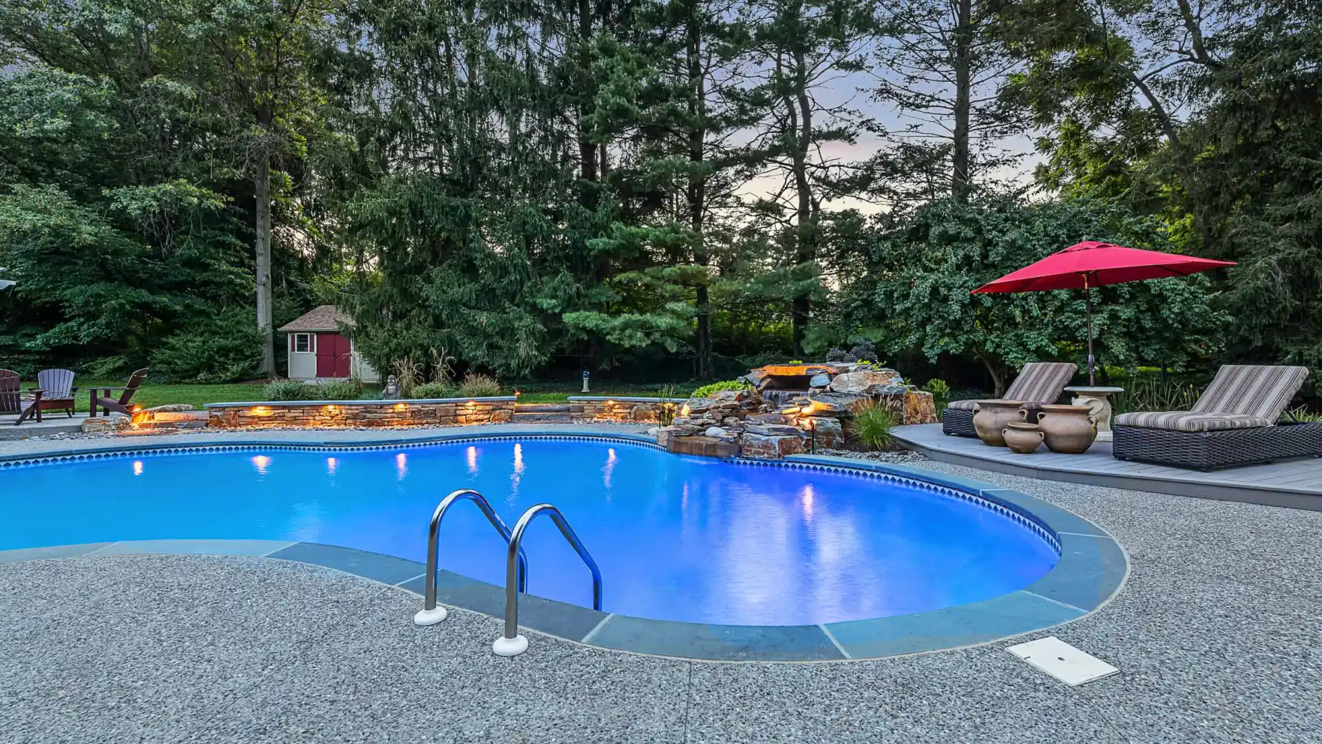 A backyard swimming pool with clear blue water is surrounded by a stone patio, showcasing impeccable outdoor living Delaware County PA appreciates. Two lounge chairs with a red umbrella sit on the right. Trees and a small red shed are in the background, with a waterfall feature cascading into the pool.