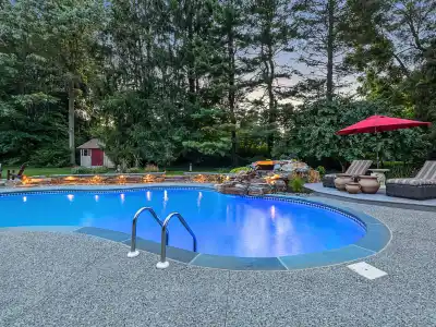 A backyard swimming pool with clear blue water is surrounded by a stone patio, showcasing impeccable outdoor living Delaware County PA appreciates. Two lounge chairs with a red umbrella sit on the right. Trees and a small red shed are in the background, with a waterfall feature cascading into the pool.