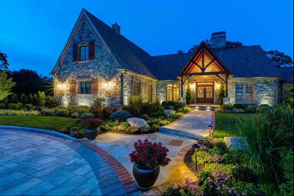 Charming stone house with peaked roof, warmly lit exterior, large front windows, and a landscaped garden with colorful flowers and shrubs at dusk. A curved driveway leads to the inviting entrance.