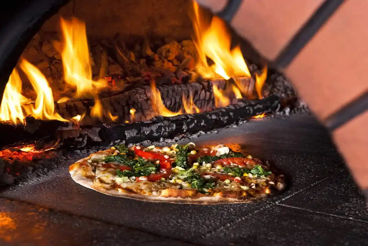 A thin-crust pizza topped with vegetables is baking inside a wood-fired oven, enhancing the outdoor living Delaware County PA ambiance. Bright flames and glowing embers add warmth to this rustic setting, perfect for enjoying moments under the stars.