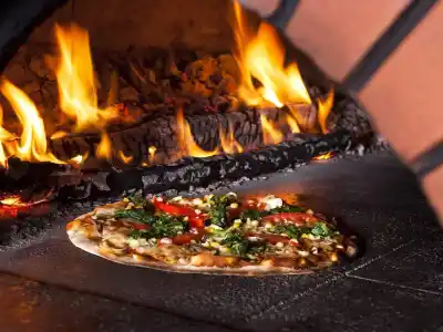 A thin-crust pizza topped with vegetables is baking inside a wood-fired oven, enhancing the outdoor living Delaware County PA ambiance. Bright flames and glowing embers add warmth to this rustic setting, perfect for enjoying moments under the stars.