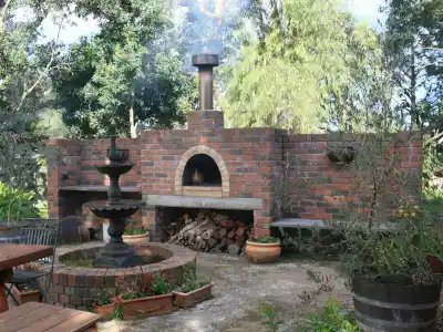 A rustic outdoor living scene in Delaware County, PA, showcases a red brick wood-fired pizza oven with logs stored underneath. A black metal tiered fountain and wooden table sit in front, surrounded by lush greenery and trees, illustrating exquisite landscaping services.