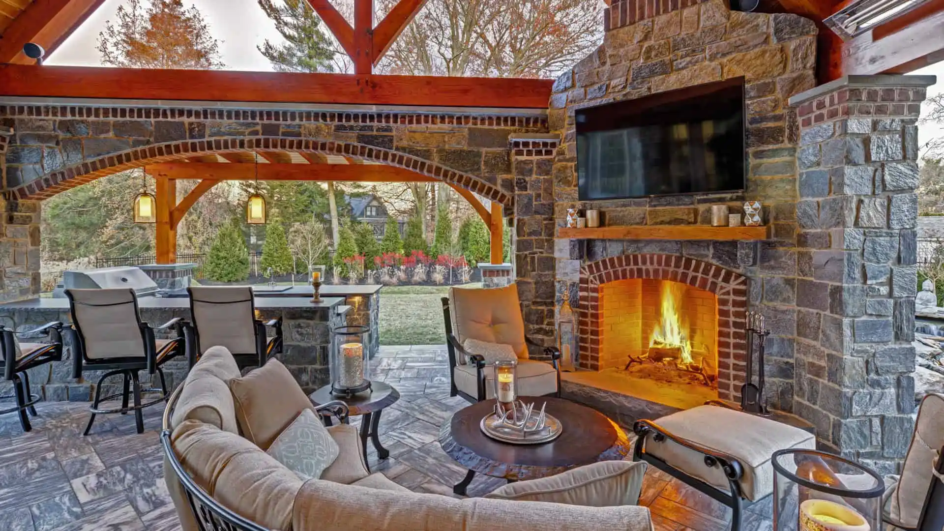 A cozy outdoor living space in Delaware County, PA features a stone fireplace with a crackling fire, cushioned seating, and a round coffee table adorned with candles. A bar area is tucked under a wooden pergola, while expert hardscaping services frame the trees and garden in the distance.