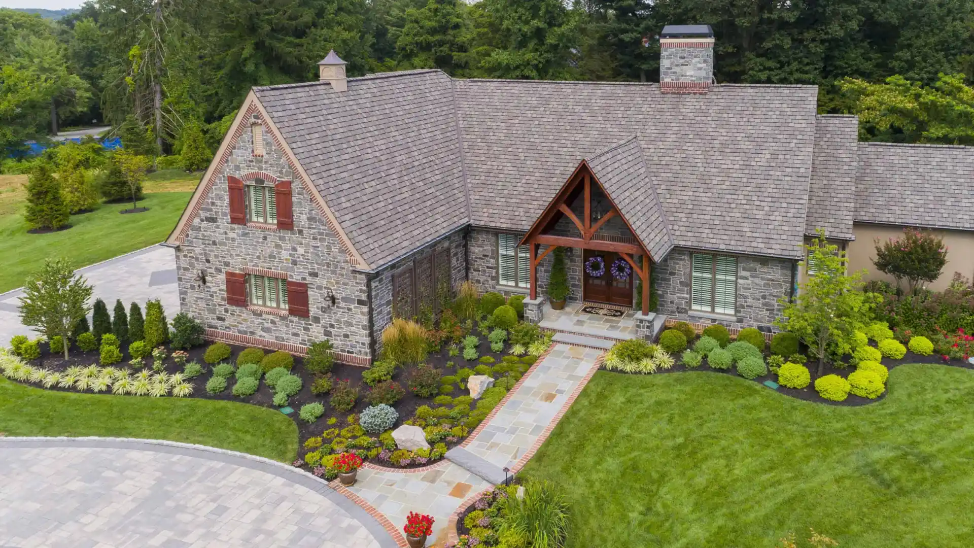 An aerial view of a stone house with a pitched roof, nestled amid exquisite outdoor living. The well-maintained garden showcases vibrant flowers, shrubs, and a curved driveway leading to the entrance. Mature trees provide a lush backdrop in Delaware County, PA.