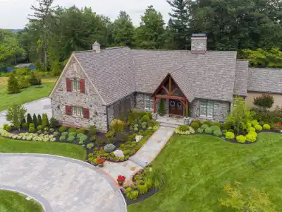 An aerial view of a stone house with a pitched roof, nestled amid exquisite outdoor living. The well-maintained garden showcases vibrant flowers, shrubs, and a curved driveway leading to the entrance. Mature trees provide a lush backdrop in Delaware County, PA.