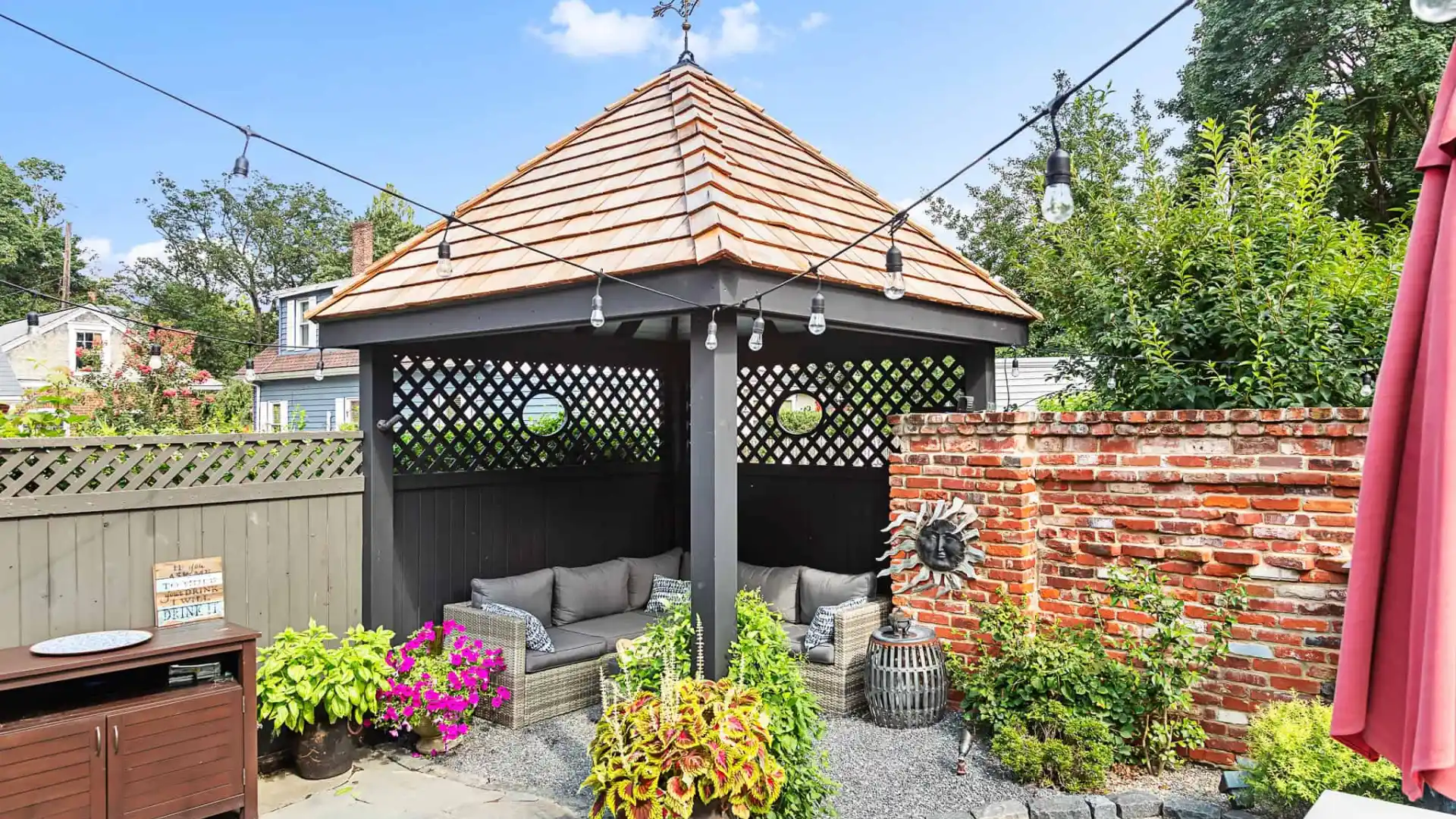 A backyard scene in Delaware County, PA, with a cozy gazebo featuring a wooden roof and lattice walls. Inside, cushioned seats await. String lights twinkle above. Vibrant potted plants and a brick wall with a sun decoration accent the area, all enhanced by expert landscaping services.