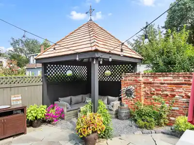 A backyard scene in Delaware County, PA, with a cozy gazebo featuring a wooden roof and lattice walls. Inside, cushioned seats await. String lights twinkle above. Vibrant potted plants and a brick wall with a sun decoration accent the area, all enhanced by expert landscaping services.