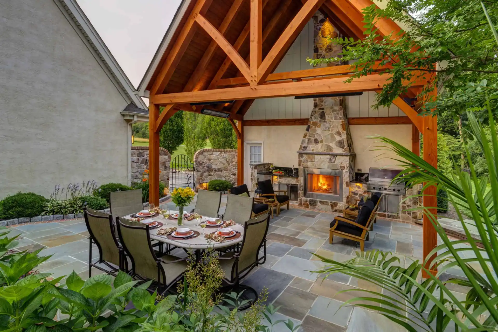 A spacious outdoor patio with a stone fireplace under a wooden pergola, featuring a dining table set for six, lounge chairs, a grill, and lush green plants surrounding the area.