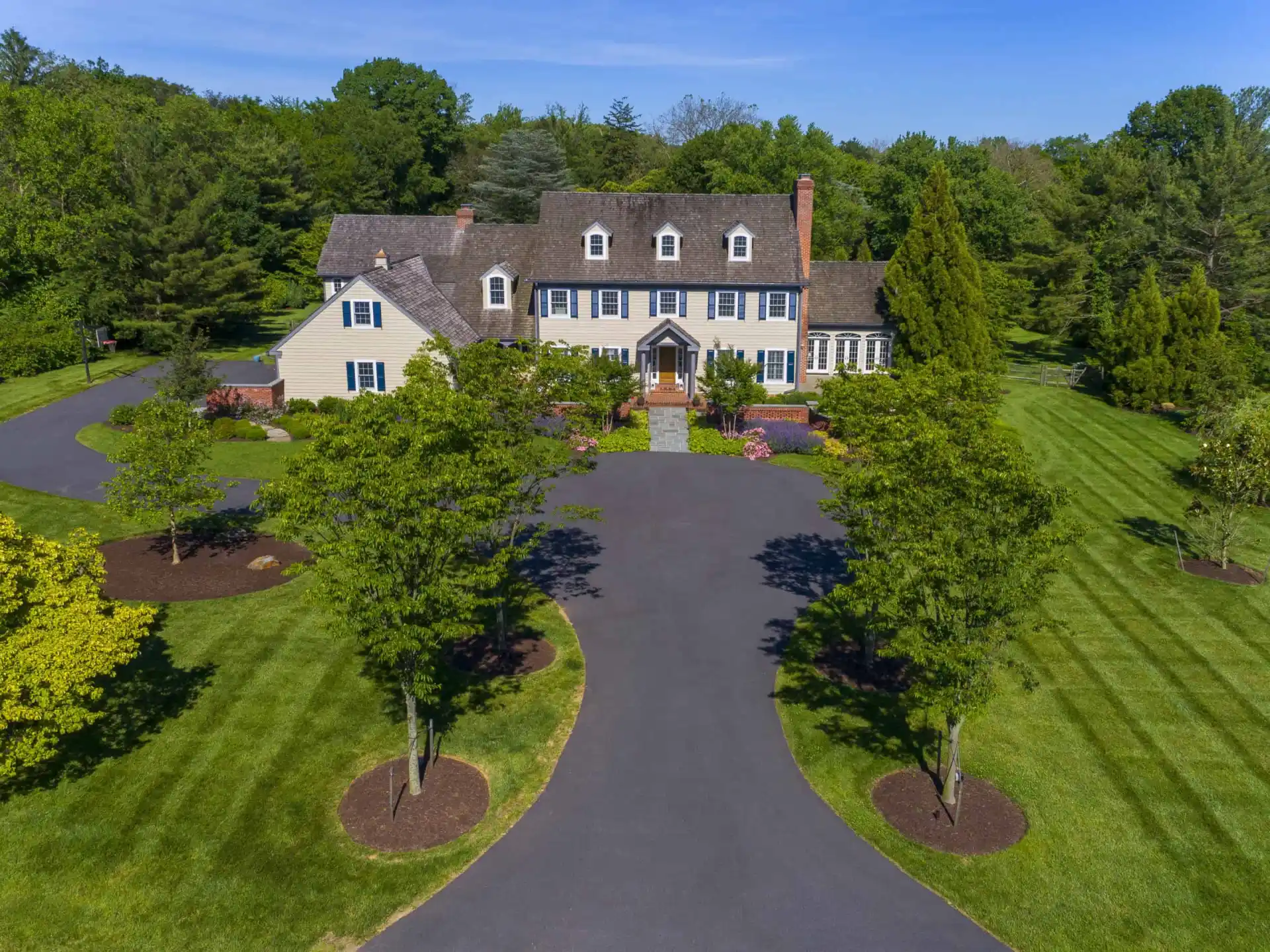 A large, two-story house with a manicured lawn, circular driveway, and landscaped trees and bushes, set against a backdrop of dense green trees under a blue sky.