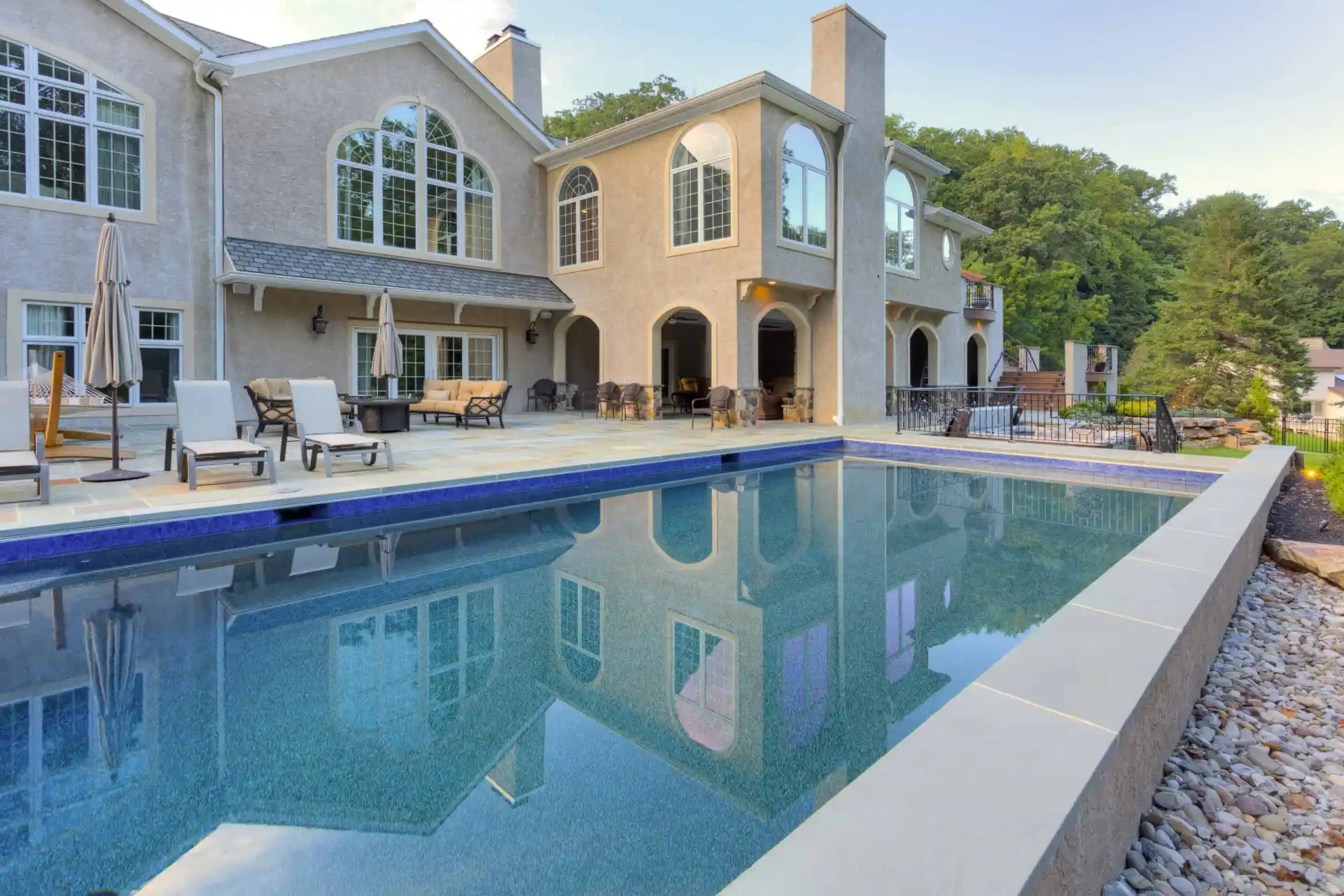 A luxurious house with large arched windows and a spacious patio is reflected in a rectangular swimming pool, surrounded by lounge chairs, umbrellas, and landscaping with rocks and trees.