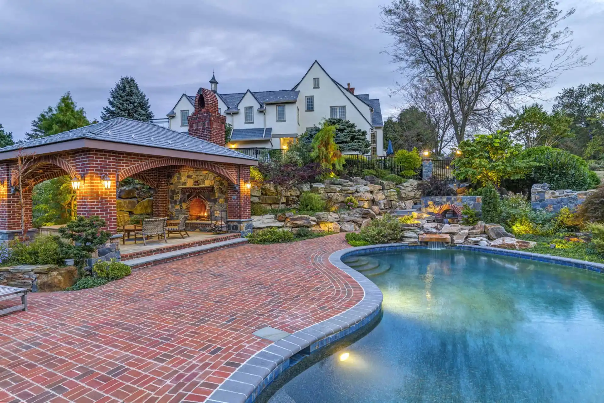 A luxurious backyard features a curved pool, a hot tub, a brick patio, a brick gazebo with a fireplace, and lush landscaping. A large, elegant house stands in the background under a cloudy sky.