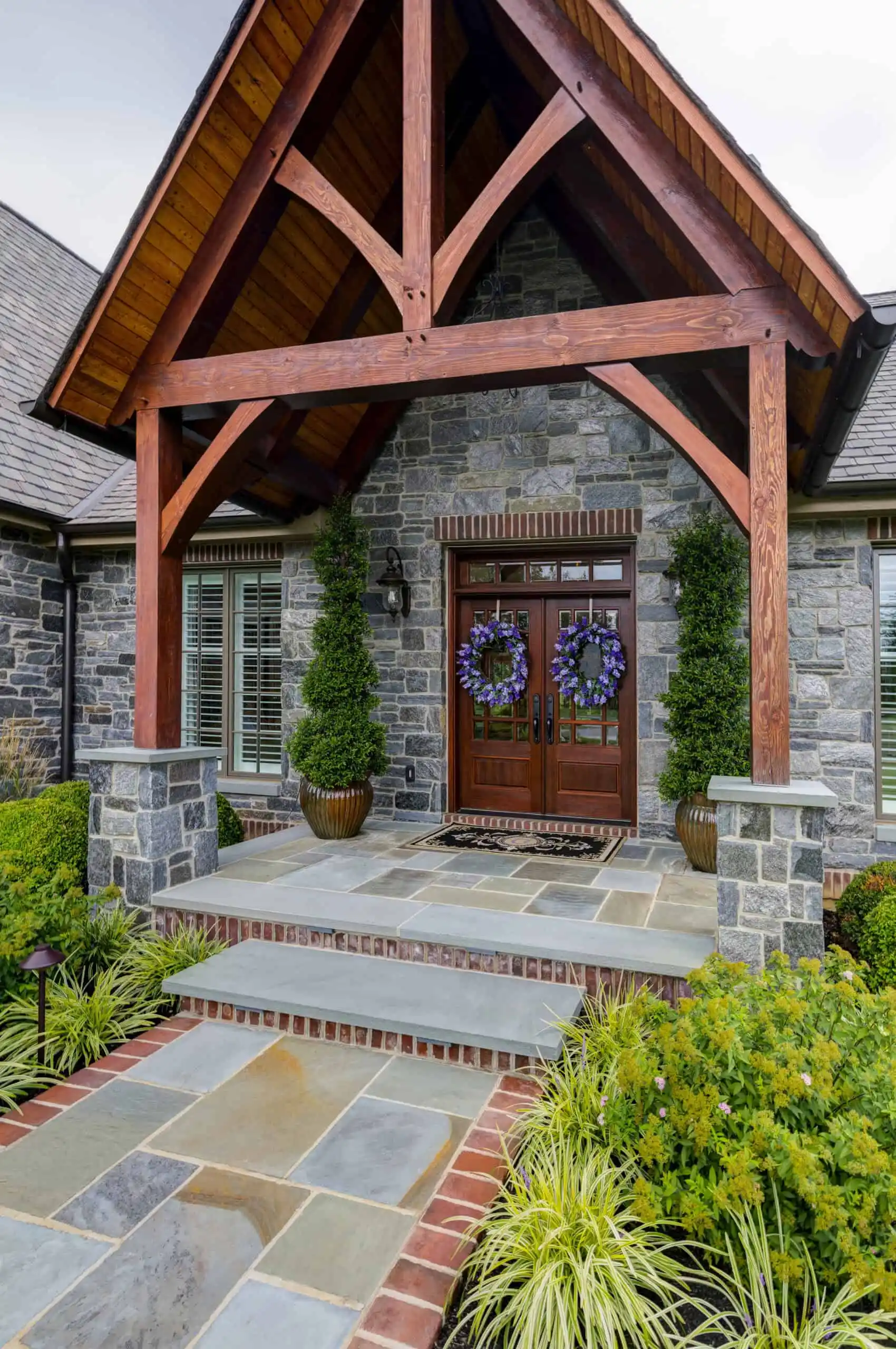 A modern stone house entrance with a wooden gabled roof, exposed beams, and large stone slabs exemplifies the artistry of hardscaping services in Delaware County, PA. The double doors boast decorative wreaths, flanked by tall potted shrubs along a walkway lined with lush greenery.