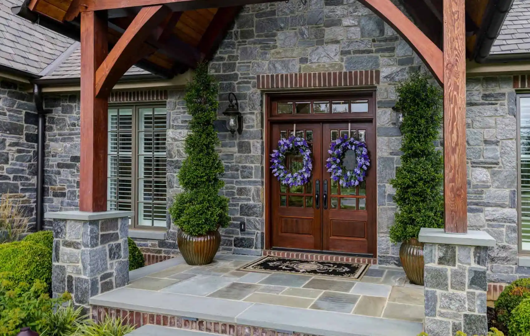 A modern stone house entrance with a wooden gabled roof, exposed beams, and large stone slabs exemplifies the artistry of hardscaping services in Delaware County, PA. The double doors boast decorative wreaths, flanked by tall potted shrubs along a walkway lined with lush greenery.