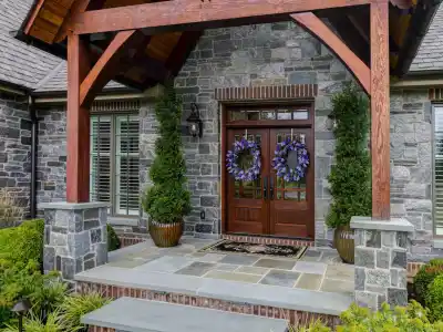 A modern stone house entrance with a wooden gabled roof, exposed beams, and large stone slabs exemplifies the artistry of hardscaping services in Delaware County, PA. The double doors boast decorative wreaths, flanked by tall potted shrubs along a walkway lined with lush greenery.