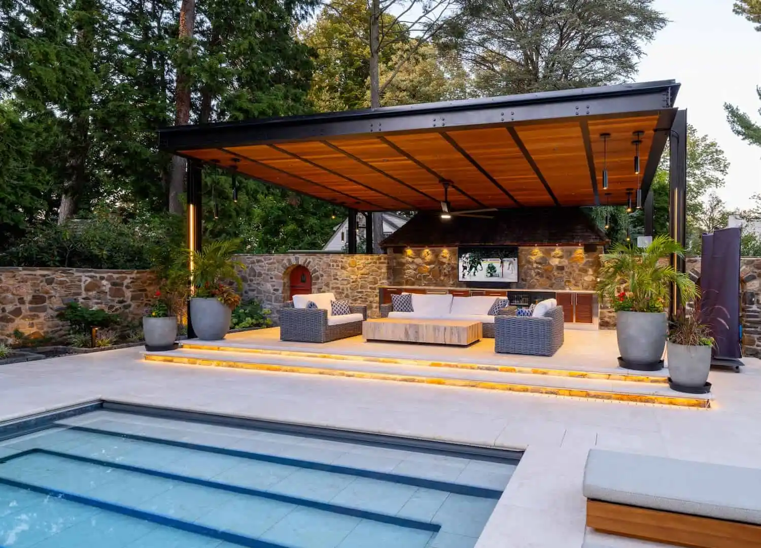Modern outdoor living space with a partially covered pergola, featuring a cozy seating area, TV, and ceiling fan. Surrounded by large potted plants and crafted through premium hardscaping services in Delaware County, PA, it overlooks a rectangular pool. Stone walls and greenery enhance the serene atmosphere.