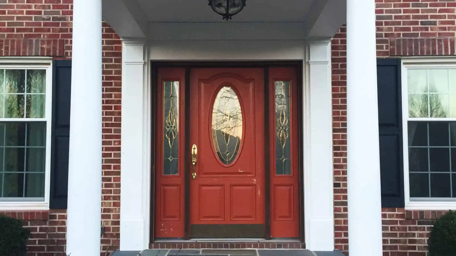 A house entrance featuring a red door with glass panels, framed by two white columns, stands as a beautiful example of outdoor living in Delaware County, PA. The red brick facade with white trim complements the steps leading up to the door and the small bare-branched tree in the foreground.