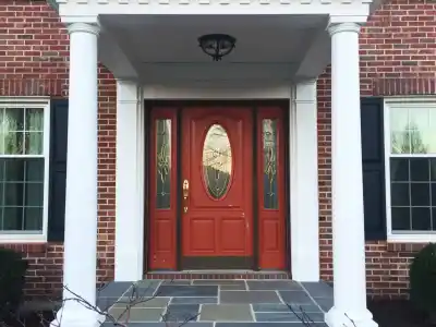 A house entrance featuring a red door with glass panels, framed by two white columns, stands as a beautiful example of outdoor living in Delaware County, PA. The red brick facade with white trim complements the steps leading up to the door and the small bare-branched tree in the foreground.
