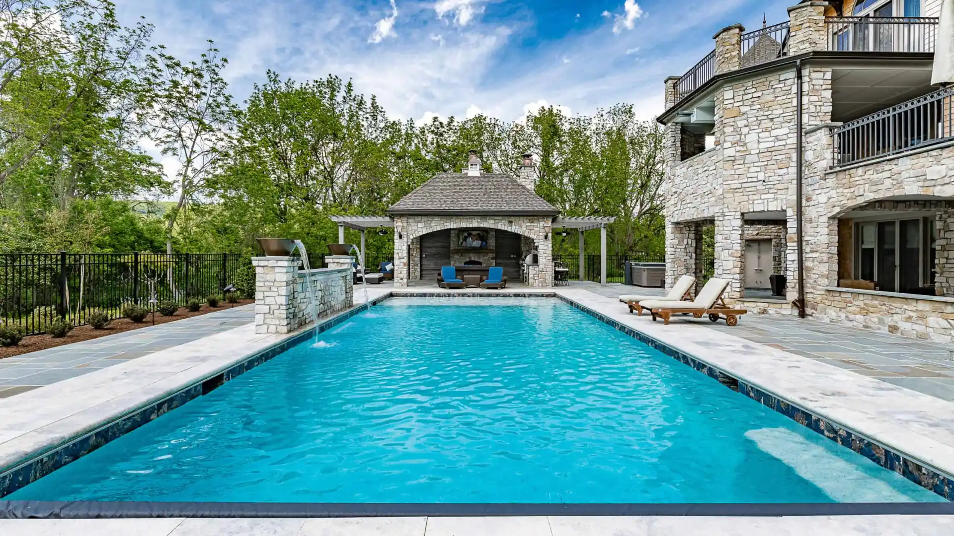Luxurious outdoor pool with clear blue water, surrounded by expertly crafted stone tiles. A charming stone building with a patio sits at the far end, framed by lush green trees. Lounge chairs grace the right side near the pool. Enjoy this example of premier hardscaping services in Delaware County, PA.