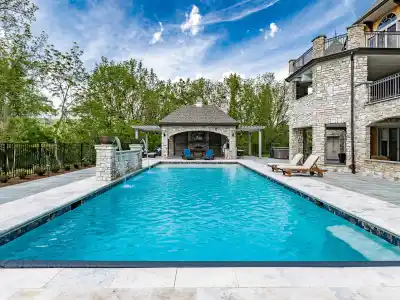 Luxurious outdoor pool with clear blue water, surrounded by expertly crafted stone tiles. A charming stone building with a patio sits at the far end, framed by lush green trees. Lounge chairs grace the right side near the pool. Enjoy this example of premier hardscaping services in Delaware County, PA.