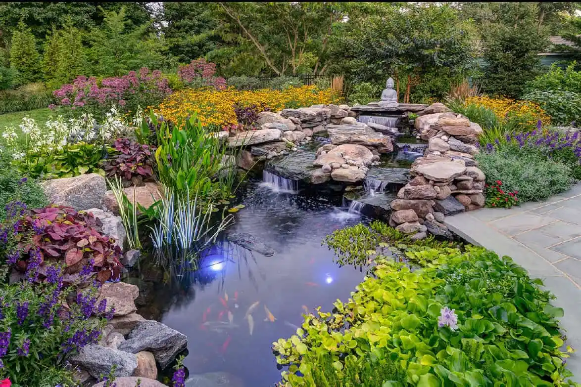 A landscaped garden pond with koi fish, surrounded by rocks, colorful flowers, lush greenery, and a small waterfall. Lights illuminate the water, and a statue sits atop the waterfall near the back.