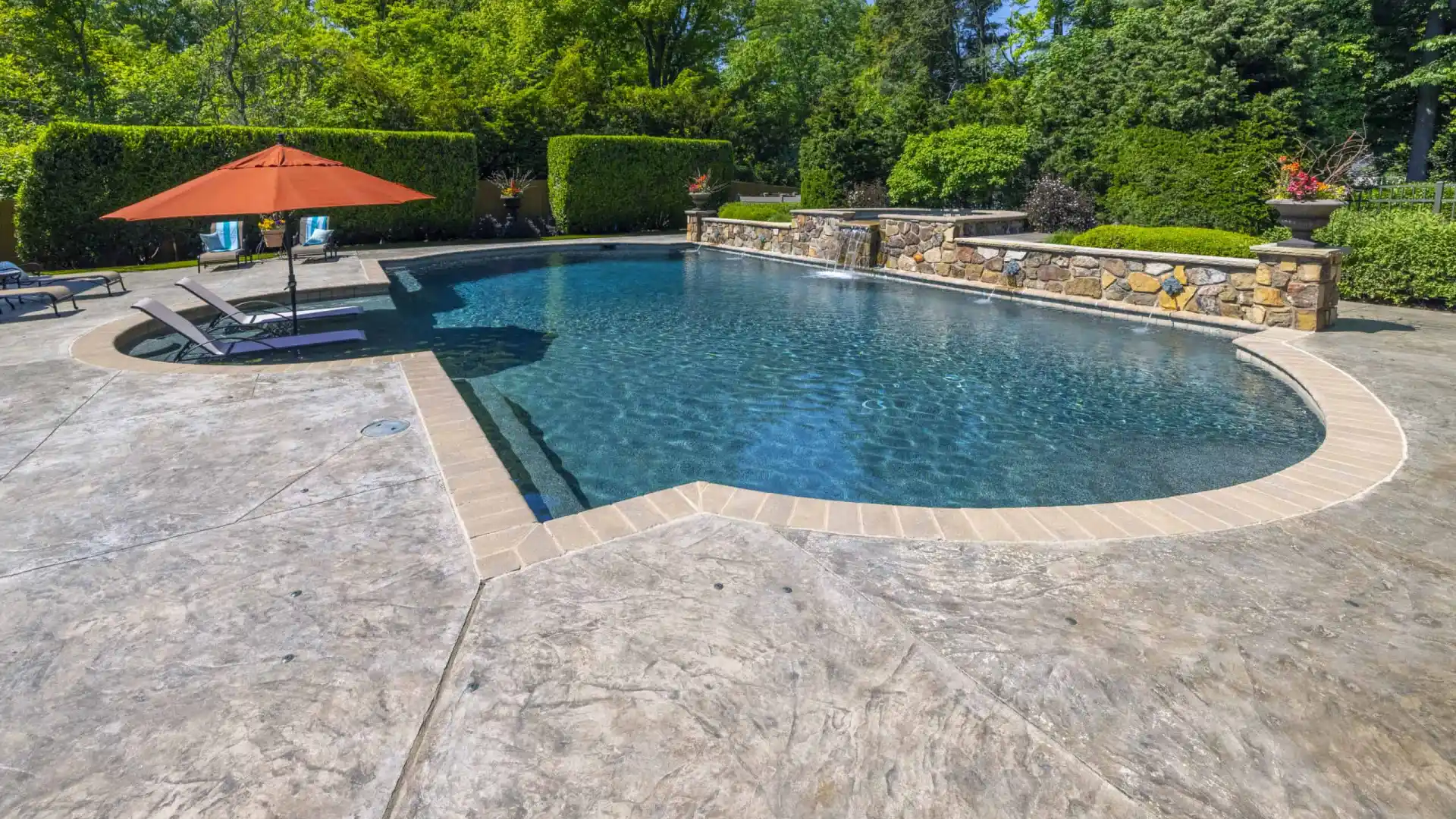 A serene backyard pool with two lounge chairs under an orange umbrella showcases exquisite landscaping services in Delaware County, PA. The textured concrete poolside area is flanked by lush green trees, while a stone wall adorned with flower pots completes the picturesque outdoor living space.