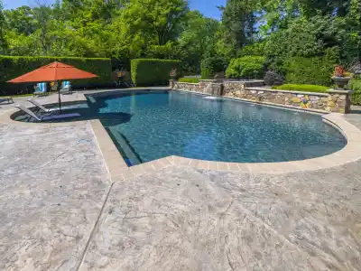 A serene backyard pool with two lounge chairs under an orange umbrella showcases exquisite landscaping services in Delaware County, PA. The textured concrete poolside area is flanked by lush green trees, while a stone wall adorned with flower pots completes the picturesque outdoor living space.
